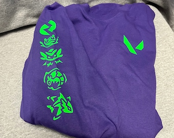 May include: A folded purple long-sleeve shirt with green graphics on the left sleeve and a green logo on the right chest. The sleeve graphics include stylized shapes. The shirt is on a gray surface.