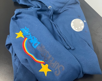 May include: A navy blue hooded sweatshirt with a rainbow and stars graphic on the left sleeve. The text "Superstar Disc Golf" is printed on the sleeve. The sweatshirt has a white moon and stars graphic on the right side.