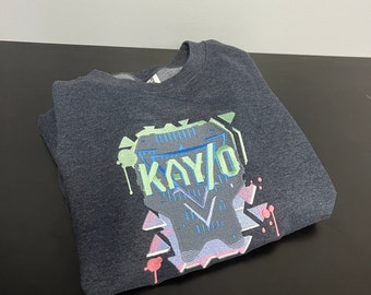 May include: A dark gray sweatshirt with a graphic print of a stylized design featuring the word "KAY/O" in green, blue, and white lettering. The design also includes geometric shapes in pink, blue, and gray.