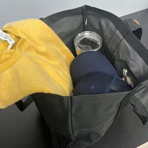 May include: A black tote bag with a yellow sweatshirt, a blue baseball cap, and a silver tumbler inside.