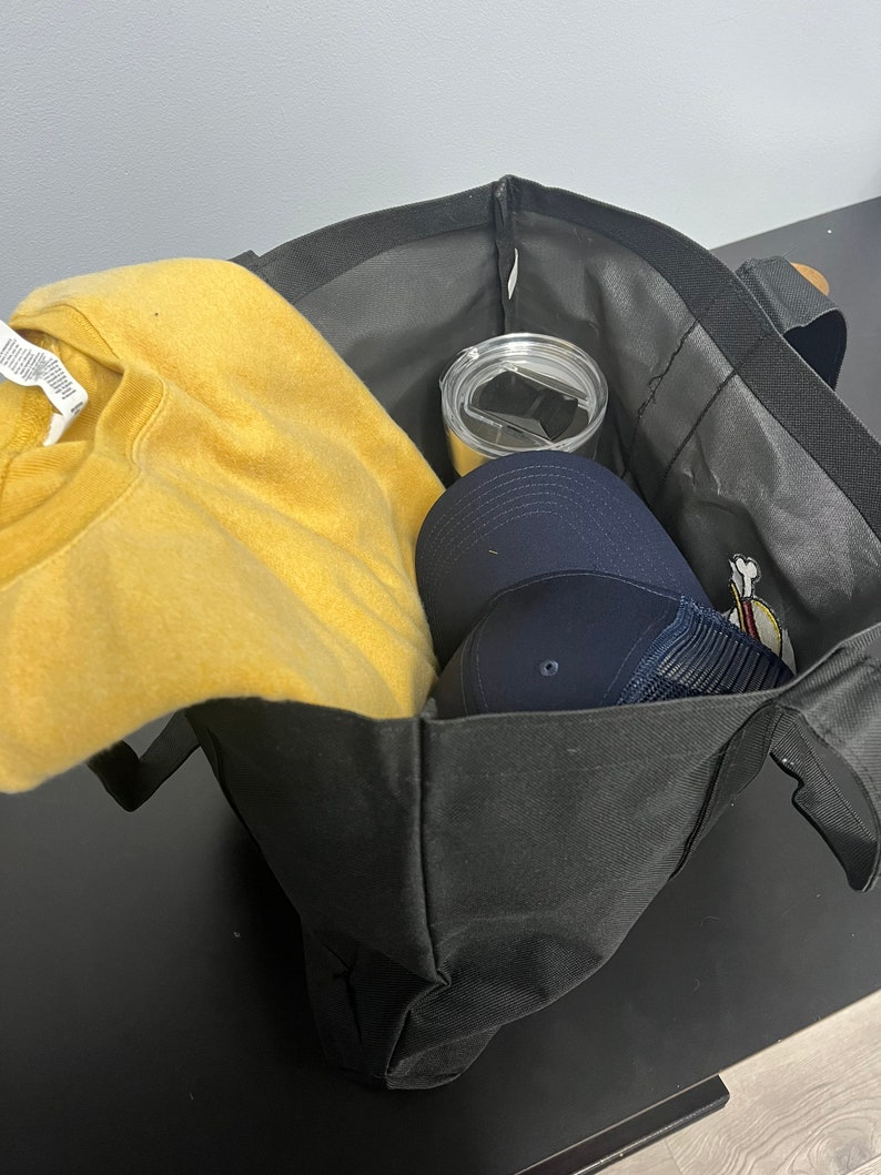 May include: A black tote bag with a yellow sweatshirt, a blue baseball cap, and a silver tumbler inside.