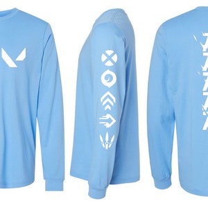 Puede incluir: Camiseta de manga larga azul claro con gráficos blancos. La parte delantera presenta un logotipo blanco, la manga tiene varios símbolos blancos y la parte trasera muestra la palabra "JET" en blanco, repetida verticalmente. Una parte superior informal y cómoda.