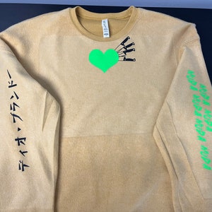 May include: A tan sweatshirt with a green heart and black knives on the front. The left sleeve has the text "タイムマシン" in black. The right sleeve has the text "MURA MURA MURA MURA" in green.