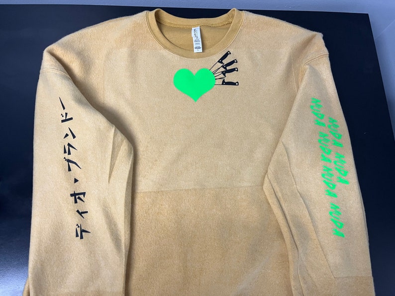 May include: A tan sweatshirt with a green heart and black knives on the front. The left sleeve has the text "タイムマシン" in black. The right sleeve has the text "MURA MURA MURA MURA" in green.