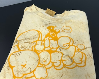 May include: A cream colored t-shirt with a distressed, faded look. The shirt features a yellow graphic of a cartoon character with a speech bubble that says "Yo!!" and the words "Fine" and "Mess" in a graffiti style.