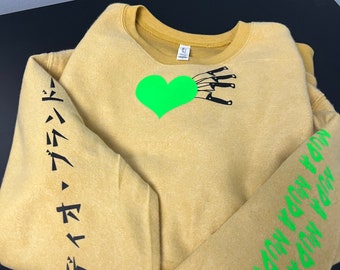 Pode incluir: Uma camisola amarela mostarda com um coração verde e facas pretas na frente. A manga esquerda tem texto japonês preto que diz "デイジー・デイジー". A manga direita tem texto verde que diz "ON THE WAY DOWN YOU WON'T FEEL SO BAD".