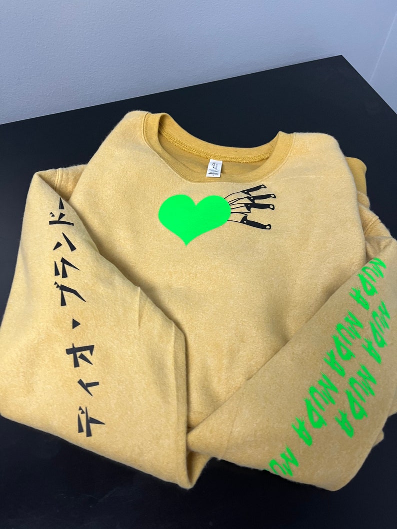 May include: A mustard yellow sweatshirt with a green heart and black knives on the front. The left sleeve has black Japanese text that reads "デイジー・デイジー". The right sleeve has green text that reads "ON THE WAY DOWN YOU WON'T FEEL SO BAD".