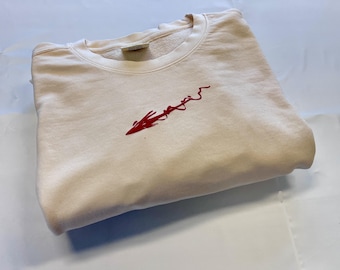 May include: A cream-colored crewneck sweatshirt featuring a long-sleeved design and a ribbed collar. Centered on the chest is an embroidered graphic in a vibrant red thread, depicting a stylized, abstract arrow-like shape with a jagged, flowing line.