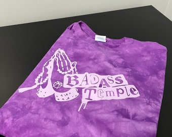 May include: Purple tie-dye t-shirt with a white graphic of praying hands holding a rosary and the text "BADASS Temple".