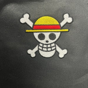 May include: White embroidered skull and crossbones with a yellow and red straw hat on a black fabric background.