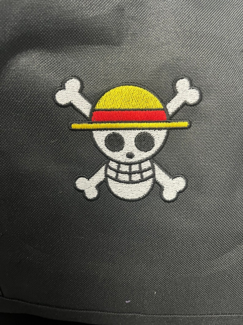 May include: White embroidered skull and crossbones with a yellow and red straw hat on a black fabric background.