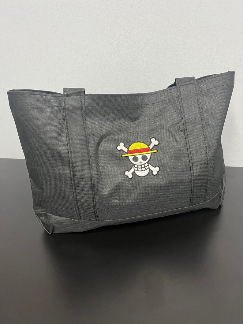May include: Black tote bag with a white skull and crossbones design. The skull is wearing a yellow and red straw hat.