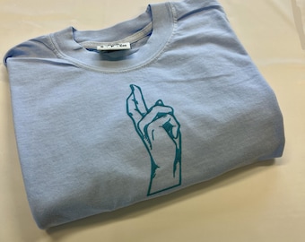 May include: A light blue crew neck t-shirt featuring a dark teal graphic print of a hand with crossed fingers on the front. The interior neck label displays the text S-P-CH. The garment is folded and laid flat, showcasing the short sleeves.