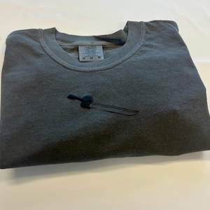 May include: A charcoal gray crew neck t-shirt featuring a small, black embroidered graphic of a sword on the chest. The inner neck label displays the text Comfort Colors and size M. The fabric has a slightly faded, washed-out appearance.