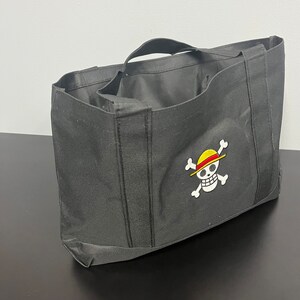 May include: Black tote bag with a white embroidered skull and crossbones design. The skull is wearing a yellow and red straw hat.