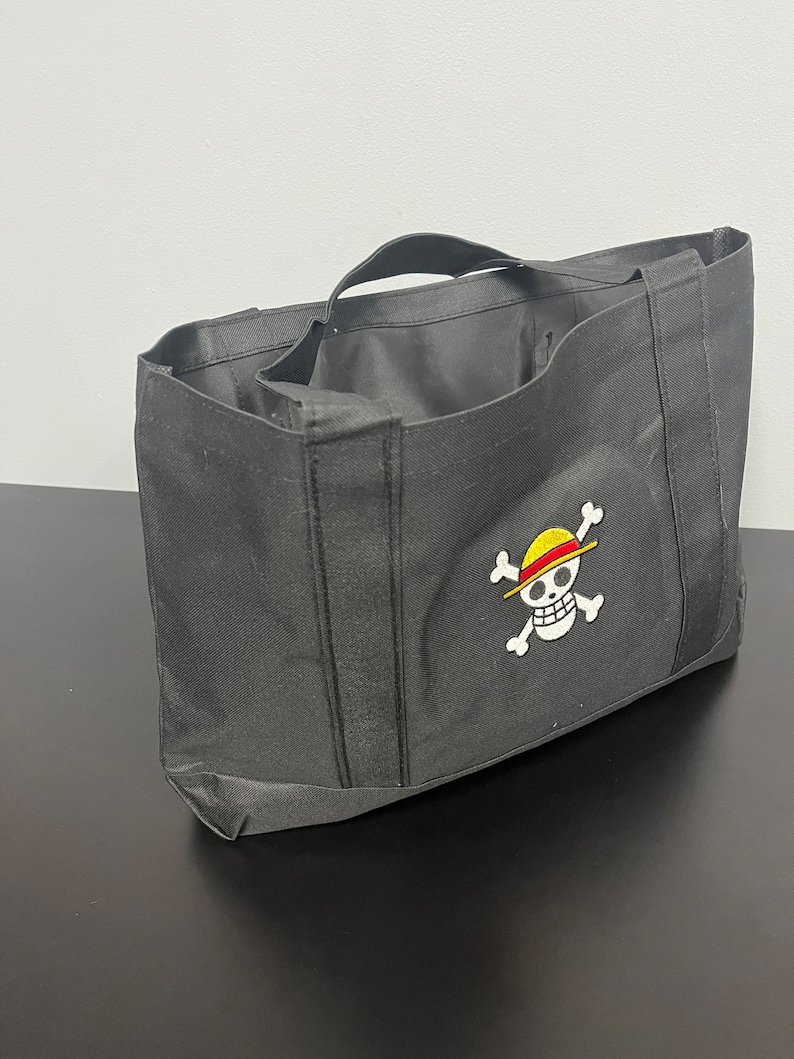 May include: Black tote bag with a white embroidered skull and crossbones design. The skull is wearing a yellow and red straw hat.