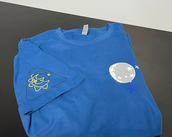 May include: A blue t-shirt with a celestial design. The shirt features a yellow sun graphic on the sleeve and a moon and star design on the front. The shirt is folded on a black surface.