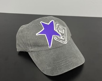 May include: A charcoal gray baseball cap featuring a large, solid purple star graphic on the front panel. Next to the star is a white, hand-drawn style rectangular emblem containing a white hand icon with a black outline.