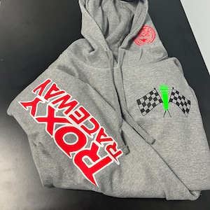 Puede incluir: Sudadera gris con capucha con un logotipo "Raceway Rox" rojo y blanco en la manga izquierda y un diseño de bandera a cuadros verde y negro en el lado derecho.