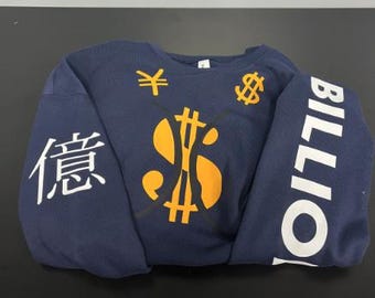 May include: A navy blue long-sleeve sweatshirt featuring a large yellow dollar sign graphic in the center, a white Japanese character on the left sleeve, and the white text BILLION printed vertically along the right sleeve with currency symbols.
