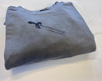 May include: A slate gray crewneck sweatshirt featuring a dark navy blue embroidered design on the chest. The embroidery depicts a sword with a detailed hilt and a long, slender blade, positioned diagonally across the center of the garment.