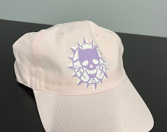 May include: A pink baseball cap with a purple skull and cat design. The design features a skull with a cat's head in place of the jawbone, surrounded by a circle of spikes.