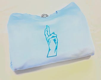 May include: A light blue crewneck sweatshirt featuring a centered graphic of a hand with crossed fingers in a darker shade of blue. The garment has a standard ribbed neckline and a visible white interior care label at the back of the collar.