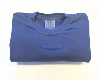 May include: A blue short-sleeve t-shirt with a crew neckline and a small embroidered barbed wire design centered on the chest. The inner neck label reads Comfort Colors, 100% cotton, and size S-P-CH. The fabric has a solid, matte blue finish.