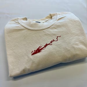 May include: A cream-colored crew neck t-shirt featuring a red embroidered graphic on the chest that resembles a stylized arrow or brushstroke. The interior collar tag displays the word COMFORT in black text on a white background.