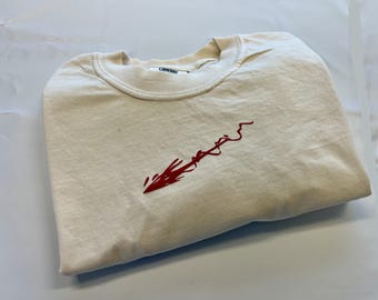 May include: A cream-colored crew neck t-shirt featuring a red embroidered graphic on the chest that resembles a stylized arrow or brushstroke. The interior collar tag displays the word COMFORT in black text on a white background.