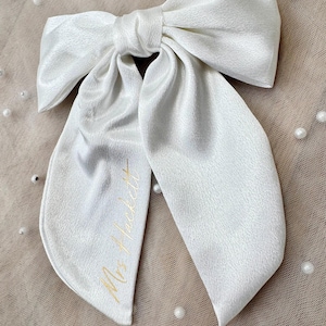 May include: A large, white satin bow with long, flowing tails. The bow is tied in the center and features the gold text "Mrs. Hackett" on one tail. The bow is likely for a special occasion, such as a wedding.