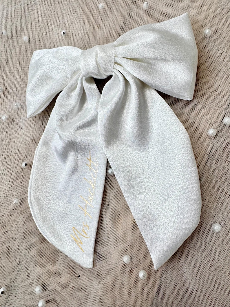 May include: A large, white satin bow with long, flowing tails. The bow is tied in the center and features the gold text "Mrs. Hackett" on one tail. The bow is likely for a special occasion, such as a wedding.
