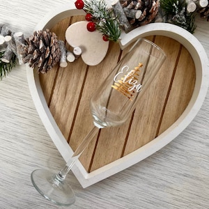 May include: A clear champagne flute with a gold Christmas tree design and the name "Eliza" in gold script. The glass is set on a wooden heart-shaped tray with white trim, decorated with pinecones and greenery.