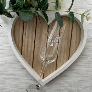 May include: A clear champagne flute with the initials "B&J" in silver script, resting on a wooden heart-shaped tray with a white border. Greenery accents the top left corner, suggesting a celebratory theme.