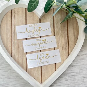 Pode incluir: Tr&ecirc;s cart&otilde;es brancos com letras douradas e um pequeno cora&ccedil;&atilde;o dourado. Os cart&otilde;es s&atilde;o etiquetados "Bride Hayley 21.05.22", "MOB  Margarita 21.05.22" e "MOG Lynn 21.05.22".