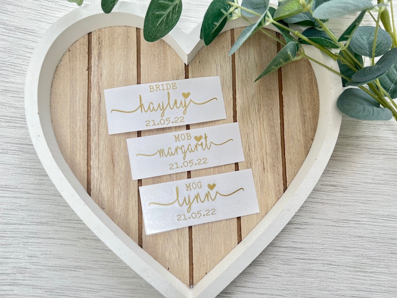 Pode incluir: Tr&ecirc;s cart&otilde;es brancos com letras douradas e um pequeno cora&ccedil;&atilde;o dourado. Os cart&otilde;es s&atilde;o etiquetados "Bride Hayley 21.05.22", "MOB  Margarita 21.05.22" e "MOG Lynn 21.05.22".