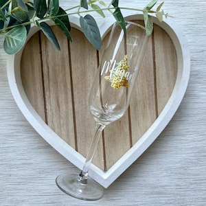 May include: A clear champagne flute with a gold glitter Christmas tree design and the name "Megan" in white script. The glass is set against a wooden heart-shaped background with a white border, and greenery.