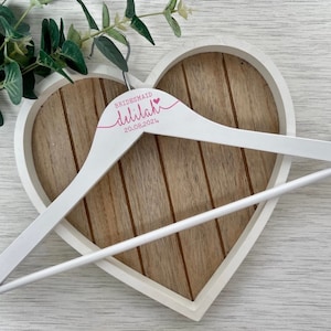 Pode incluir: Cabide de madeira branco com o texto "BRIDESMAID  dillilah 20.08.2024" em letras cor-de-rosa. O cabide est&aacute; numa bandeja de madeira em forma de cora&ccedil;&atilde;o.