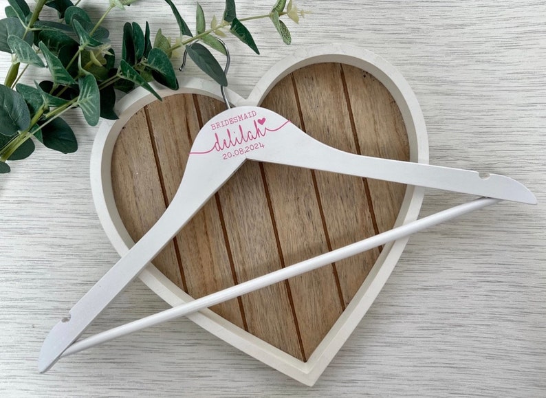 Pode incluir: Cabide de madeira branco com o texto "BRIDESMAID  dillilah 20.08.2024" em letras cor-de-rosa. O cabide est&aacute; numa bandeja de madeira em forma de cora&ccedil;&atilde;o.