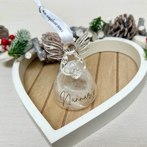 May include: A clear glass angel ornament with feathery filling, resting in a white heart-shaped wooden tray. The angel has silver wings and a ribbon that reads "in angels are near". The ornament is personalized with the name "Nannar".