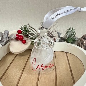 May include: A clear glass angel ornament with white feathers inside and the name "Carmen" in red script. The ornament has a white ribbon with the text "Feathers appear when" and is surrounded by decorative elements.