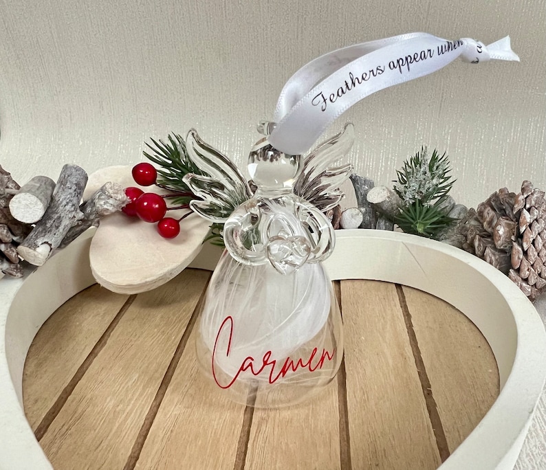 May include: A clear glass angel ornament with white feathers inside and the name "Carmen" in red script. The ornament has a white ribbon with the text "Feathers appear when" and is surrounded by decorative elements.