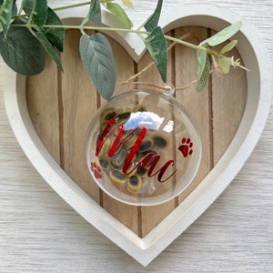 May include: A clear, round ornament filled with small, tan and brown treats. The ornament has the name "Mac" in red script, along with two red paw prints. The ornament hangs from a twine string, set against a wooden heart-shaped background.