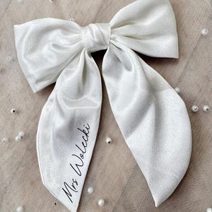 May include: A large, white satin bow with long, flowing tails. The bow is tied in the center and features the text "Mrs. Wolecki" in a delicate script on one tail. The bow is likely for a special occasion.