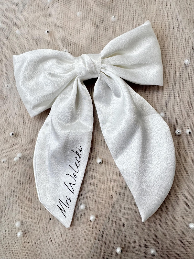 May include: A large, white satin bow with long, flowing tails. The bow is tied in the center and features the text "Mrs. Wolecki" in a delicate script on one tail. The bow is likely for a special occasion.