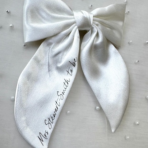 May include: A large, white satin bow with long, flowing tails. The bow is tied in the center and features the text "Mrs Stewart-Smith to be" in a delicate script. Perfect for bridal or wedding events.