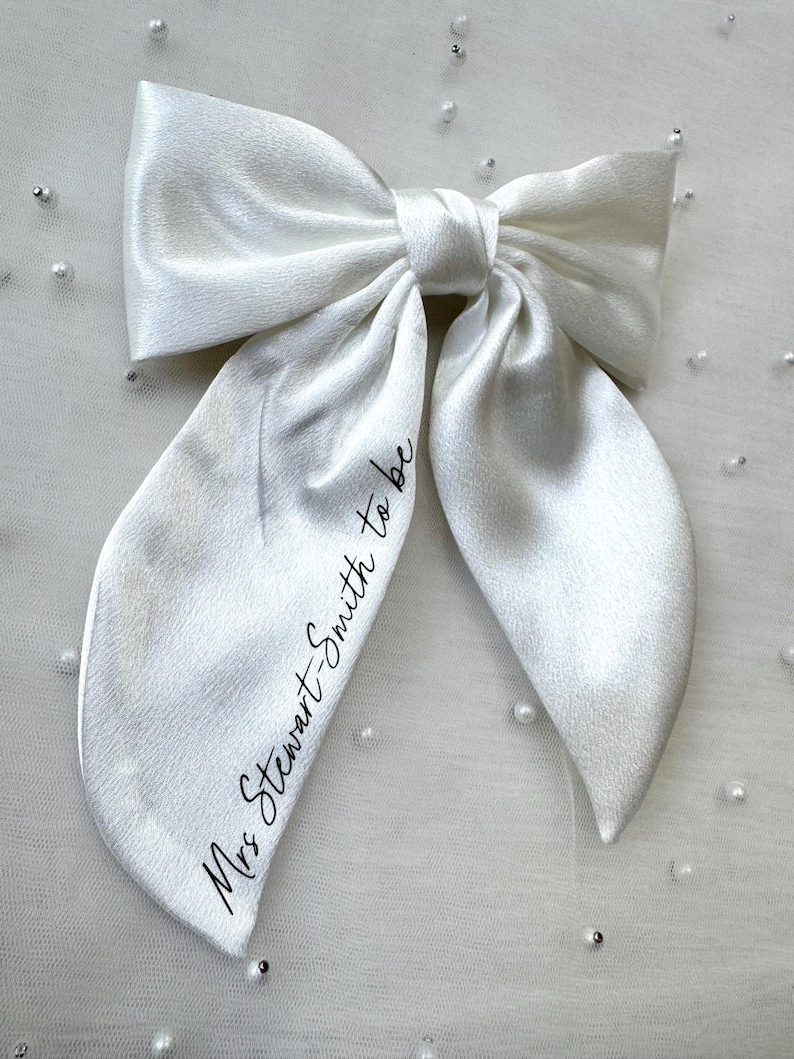May include: A large, white satin bow with long, flowing tails. The bow is tied in the center and features the text "Mrs Stewart-Smith to be" in a delicate script. Perfect for bridal or wedding events.