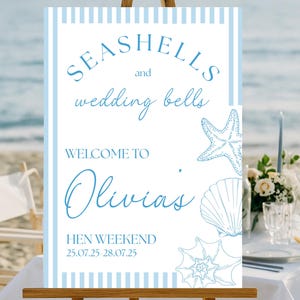 Puede incluir: Un letrero azul claro y blanco sobre un caballete de madera, con las palabras "Seashells and wedding bells" y "Welcome to Olivia's Hen Weekend". El letrero tiene ilustraciones de conchas marinas y estrellas de mar.