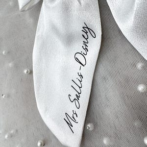 May include: Close-up of a white satin bow with the text "Mrs Sallis-Disney" in black script. The bow is set against a backdrop of white tulle dotted with small, white pearls, creating a delicate and elegant aesthetic.