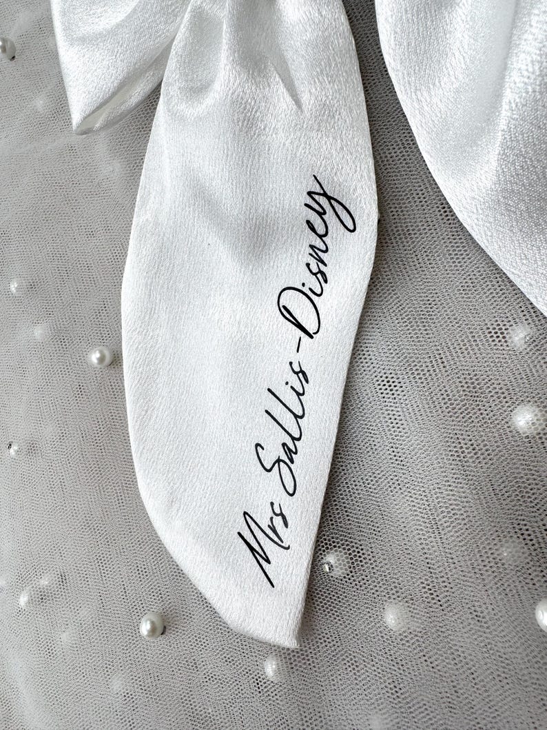 May include: Close-up of a white satin bow with the text "Mrs Sallis-Disney" in black script. The bow is set against a backdrop of white tulle dotted with small, white pearls, creating a delicate and elegant aesthetic.